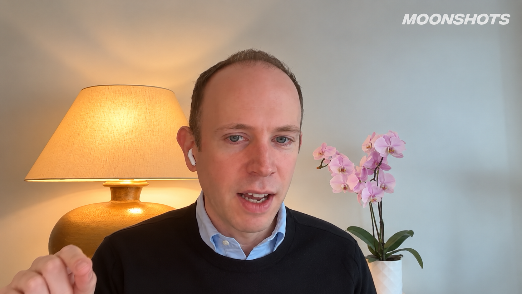 Alex Wissner-Gross gestures in a black sweater and pale blue shirt between a warm table lamp and pink orchids in a white planter. MOONSHOTS appears at the upper right.